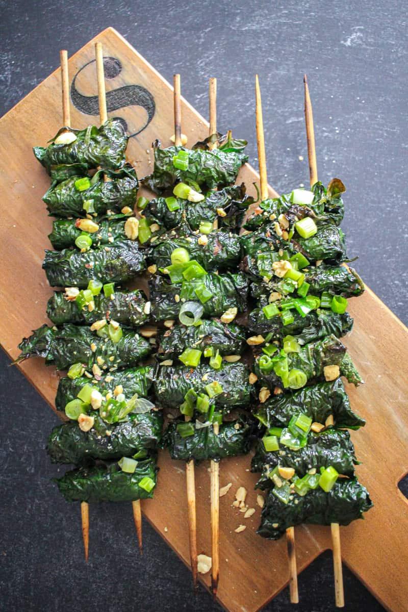 A Birds eye view of Three skewers of betel leaf wrapped beef (Bò Cuốn Lá Lốt) on a wooden board, topped with crushed peanuts and scallion oil.